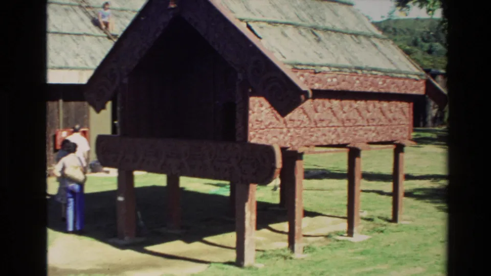 1981 New Zealand frame showing a small raised carved structure in front of a larger Maori building, with visitors on the grass.