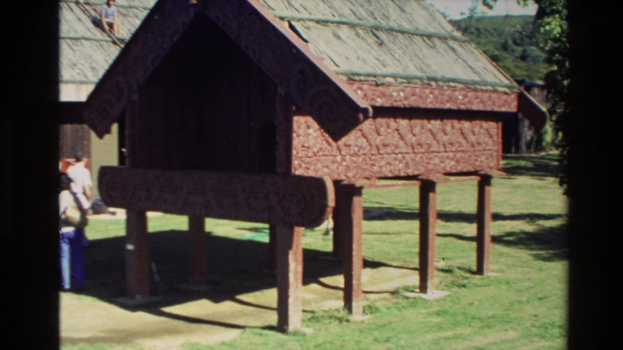 Raised red carved meeting-house structure on grass with visitors standing nearby.
