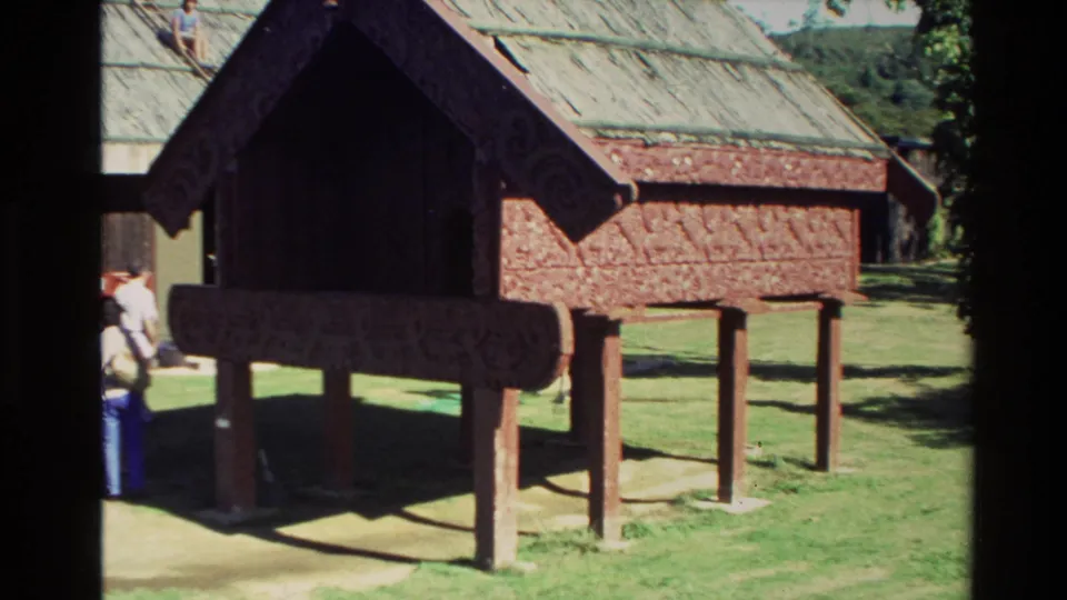 1981 New Zealand home-movie frame showing a raised Māori meeting-house structure with red carved panels and visitors at the side.