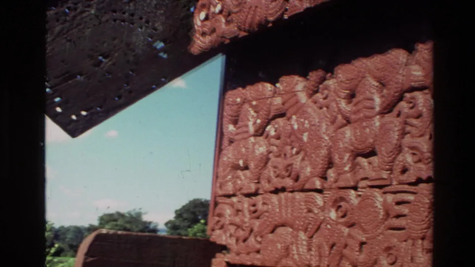 Close 8mm frame of red carved Māori architectural panels in New Zealand, with a dark roof edge and sky visible.