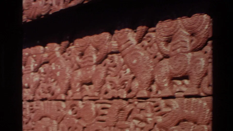 Tight 1981 New Zealand home-movie detail of red carved timber panels with repeated stylized forms and weathered texture.