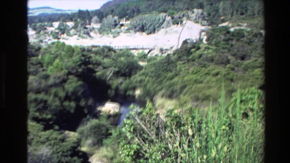 1981 New Zealand 8mm frame looking over a green river valley with dense vegetation, pale valley floor, and distant hills.