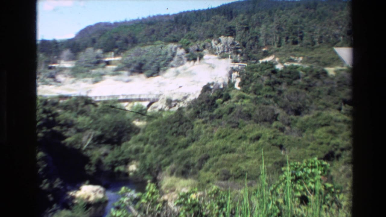 1981 New Zealand 8mm frame showing a green hillside, pale geothermal terrain, a small bridge or walkway, and dark vertical window framing.