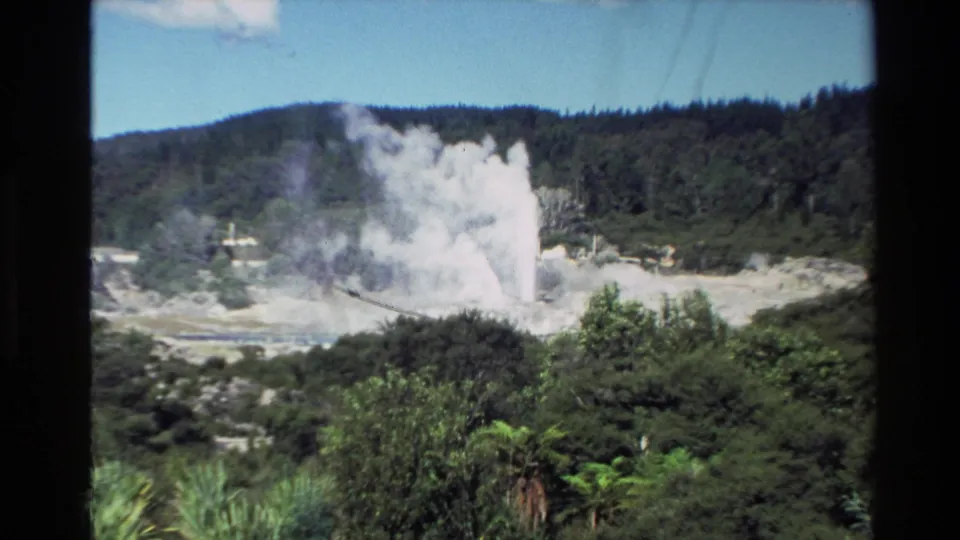 1981 New Zealand 8mm frame showing a white steam plume rising from a geothermal area, with shrubs and forested hills in the background.