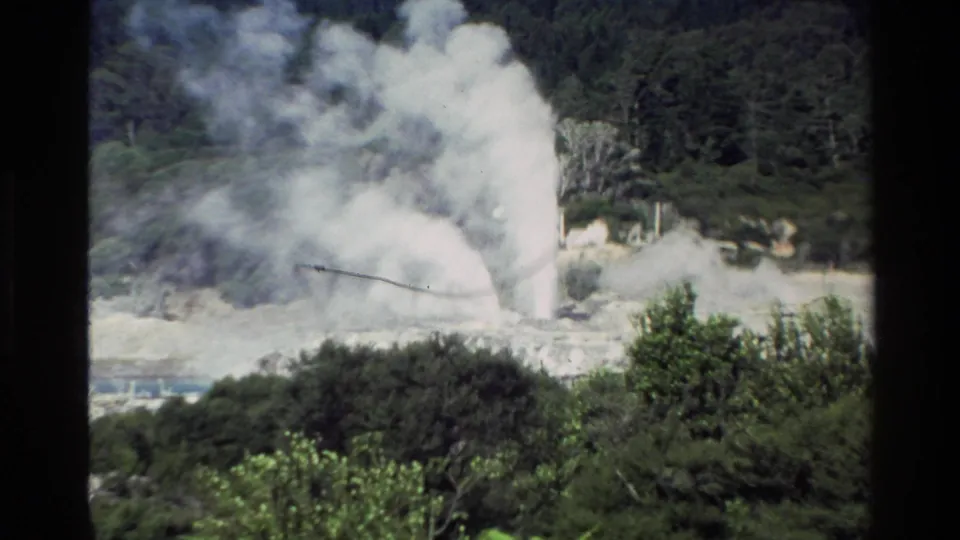 1981 New Zealand 8mm frame with a dense geothermal steam plume spreading upward and sideways over shrubs, pale ground, and a wooded slope.
