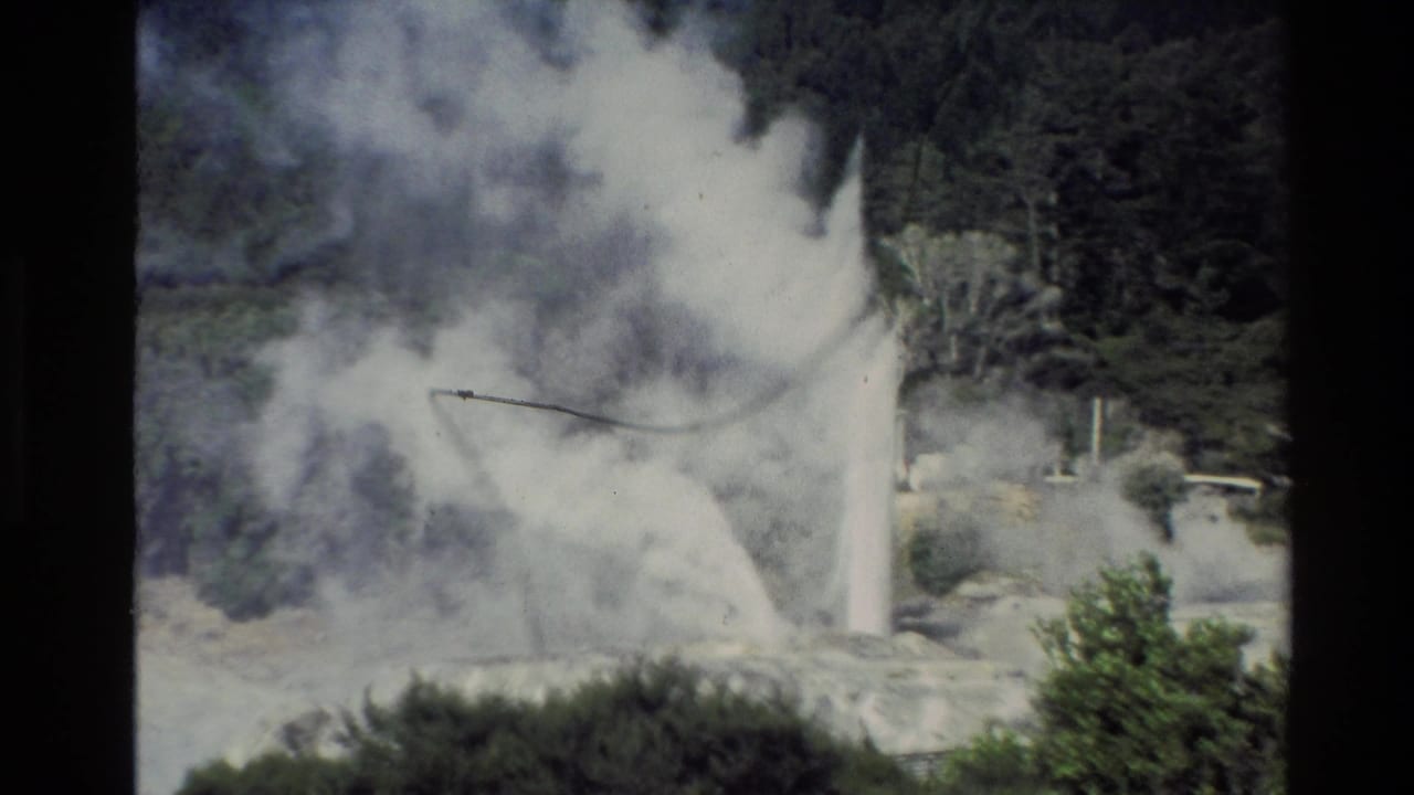 A vintage 8mm view of a New Zealand geyser eruption, with white steam spreading across pale rocks and trees in the background.