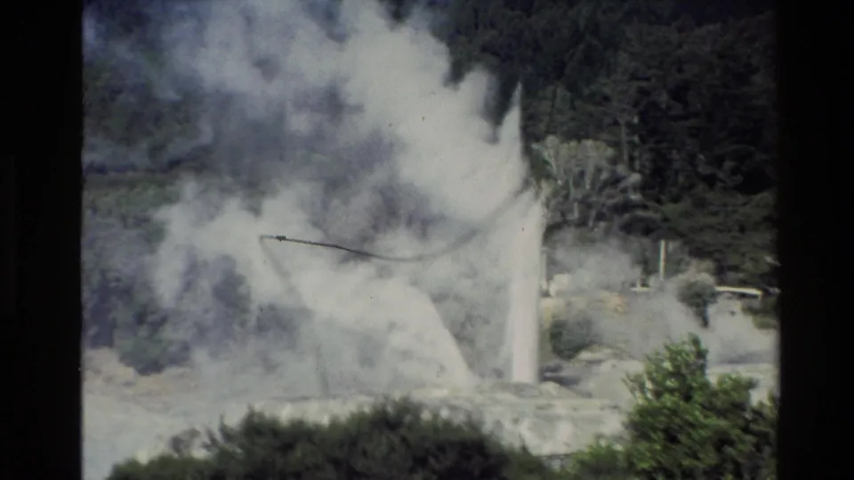 A vintage 8mm view of a New Zealand geyser eruption, with white steam spreading across pale rocks and trees in the background.