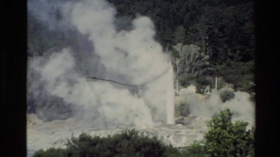 The 1981 New Zealand home-movie frame shows dense steam from a geyser eruption drifting over shrubs and pale geothermal rock.