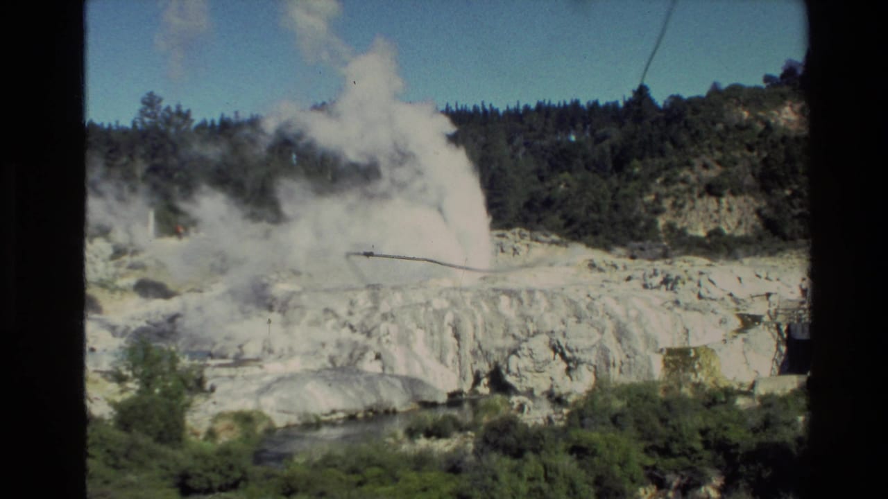 1981 New Zealand 8mm frame showing a geyser or hot spring eruption sending steam above pale mineral terraces and nearby greenery.