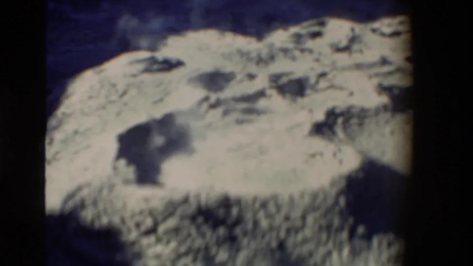 1981 New Zealand close-up of a pale geothermal mud-pot surface, with dark wet openings and rough raised rims under strong sunlight.