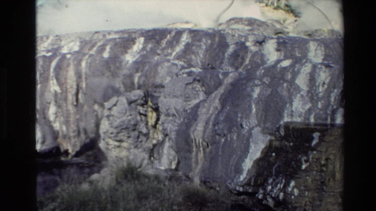 1981 New Zealand 8mm frame of a mineral-streaked cliff face with layered rock and a dark opening or shadowed recess below.