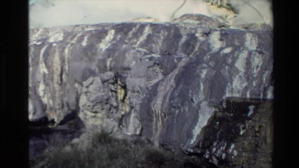 1981 New Zealand 8mm frame of a mineral-streaked cliff face with layered rock and a dark opening or shadowed recess below.