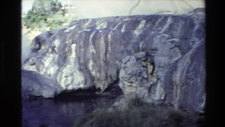 1981 New Zealand 8mm frame showing a pale mineral cliff beside dark water, with grass in the foreground and layered rock above.