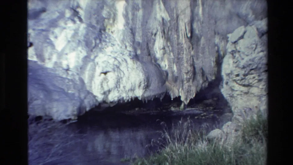 1981 New Zealand 8mm frame showing a dark cave-like recess under a mineral-streaked cliff, with water and grass at the base.