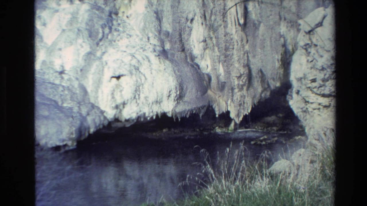 1981 New Zealand 8mm frame showing a pale mineral-encrusted rock wall, dark hot pool, grasses, and steam in a geothermal area.