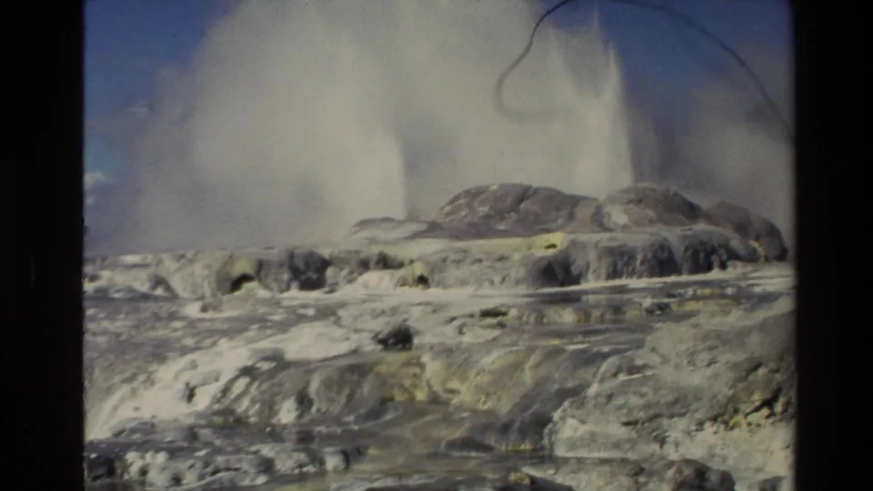 1981 New Zealand 8mm frame with a large steam plume rising from pale geothermal terraces and rocky deposits beneath blue sky.