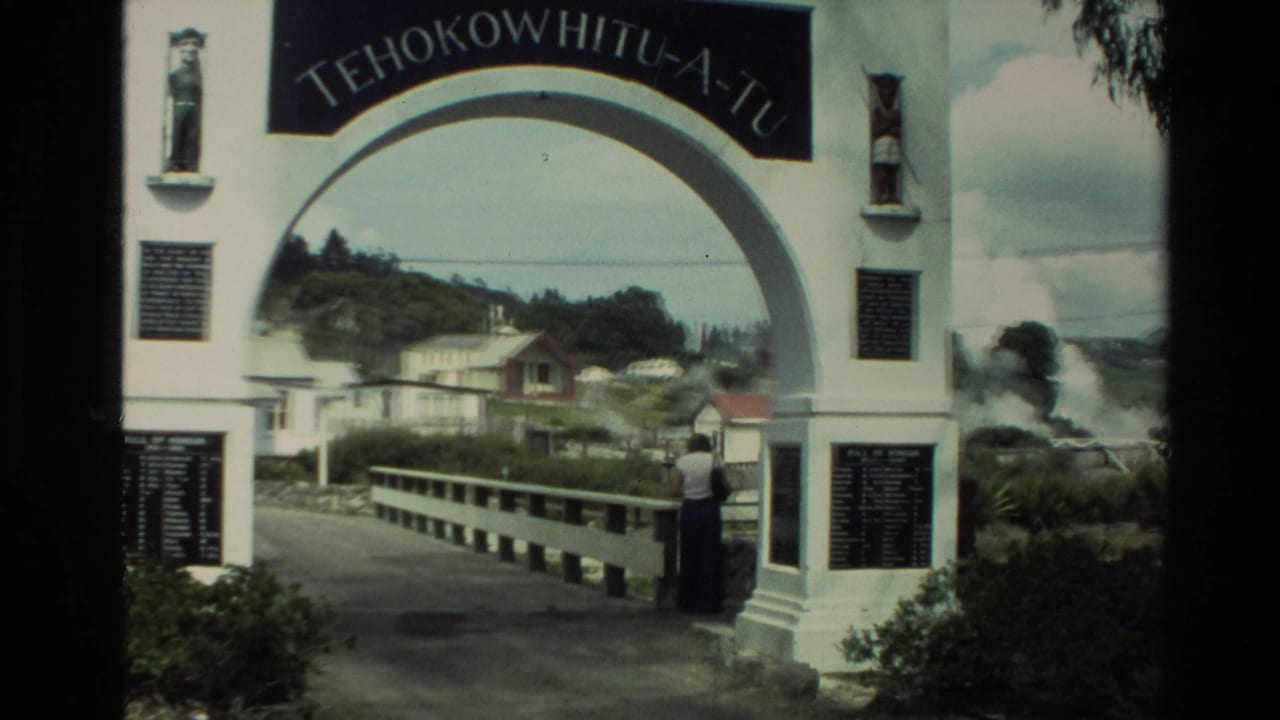White archway entrance with plaques, bridge railing, houses, hills, and a visitor.