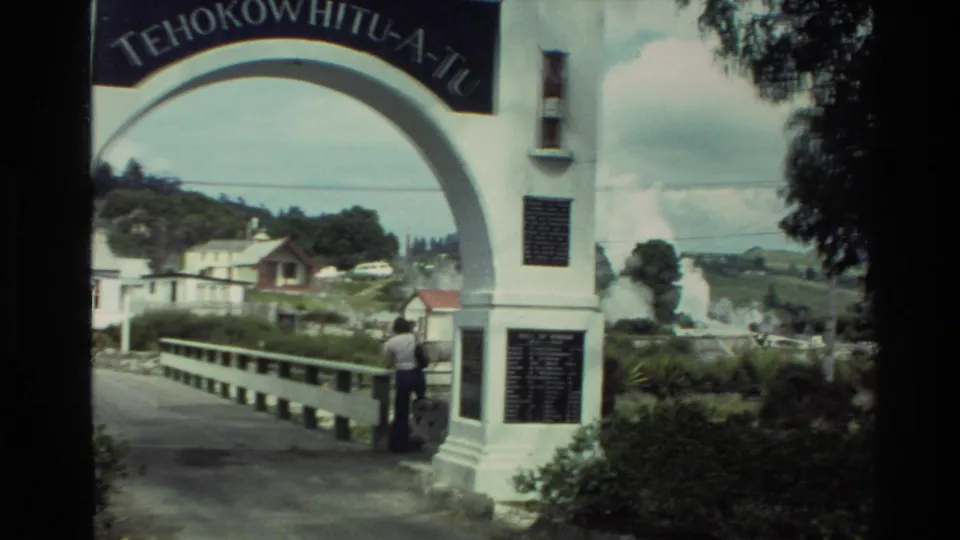 1981 New Zealand 8mm frame of a visitor beside the Te Hokowhitu-a-Tu archway, with settlement and geothermal steam beyond.