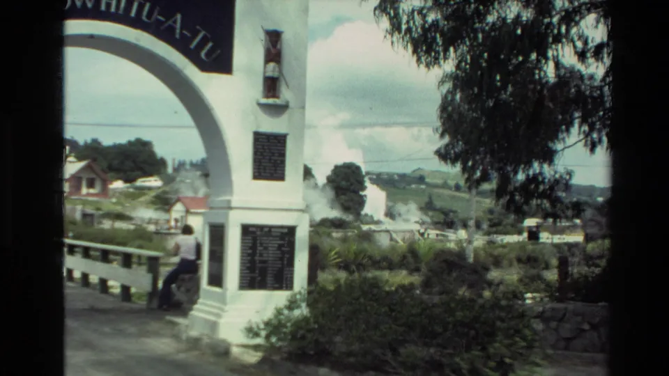 1981 New Zealand frame showing part of the Te Hokowhitu-a-Tu archway, plaques, vegetation, houses, hills, and geothermal steam.
