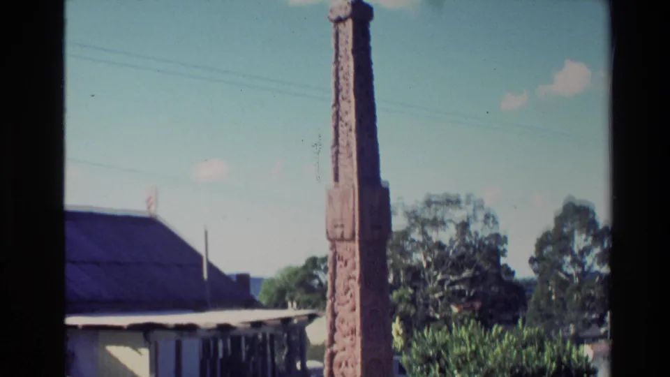 1981 New Zealand 8mm view of a tall carved monument rising above a small church building and nearby trees.