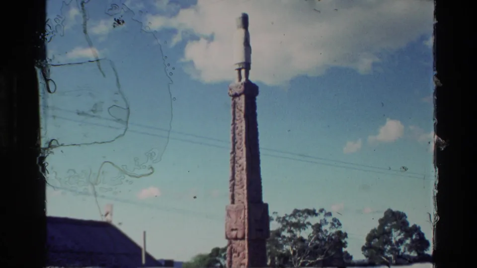 1981 New Zealand frame showing a tall carved monument against blue sky, with trees and a low roofline below.