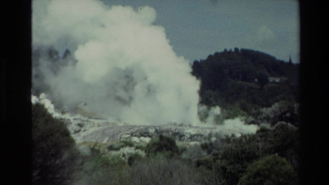 Large white steam plume rising from a rocky geothermal area with green hills behind it.