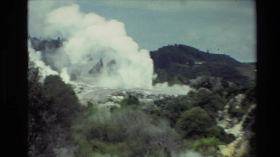 1981 New Zealand 8mm frame with white steam and darker smoke rising from a mineral-streaked geothermal area beside green vegetation.