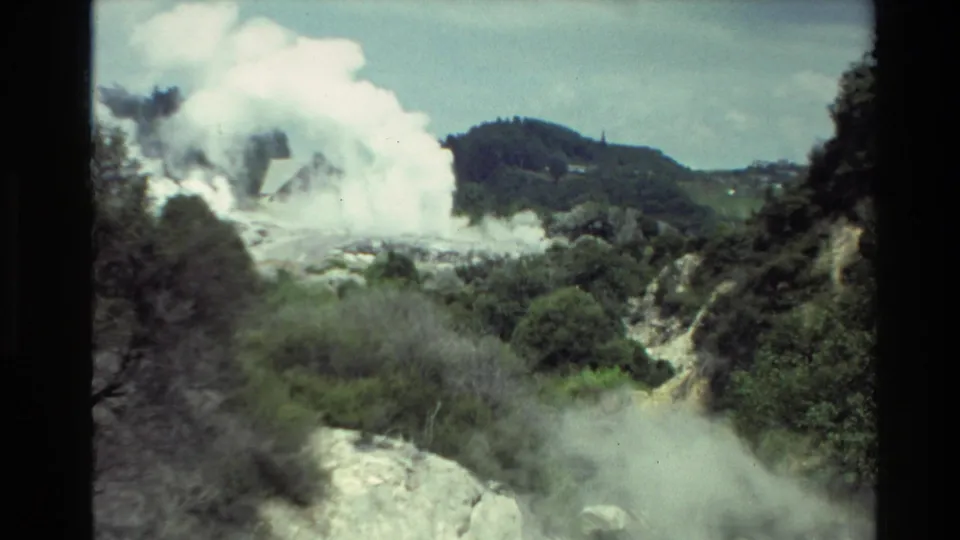 1981 New Zealand 8mm frame widening to show steam plumes across pale geothermal rock, with shrubs and rounded green hills beyond.