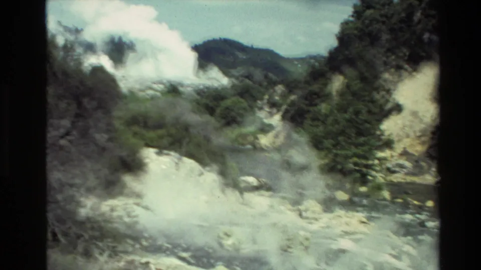 1981 New Zealand 8mm frame looking across a steaming rocky ravine toward a larger geothermal plume and green hillside.