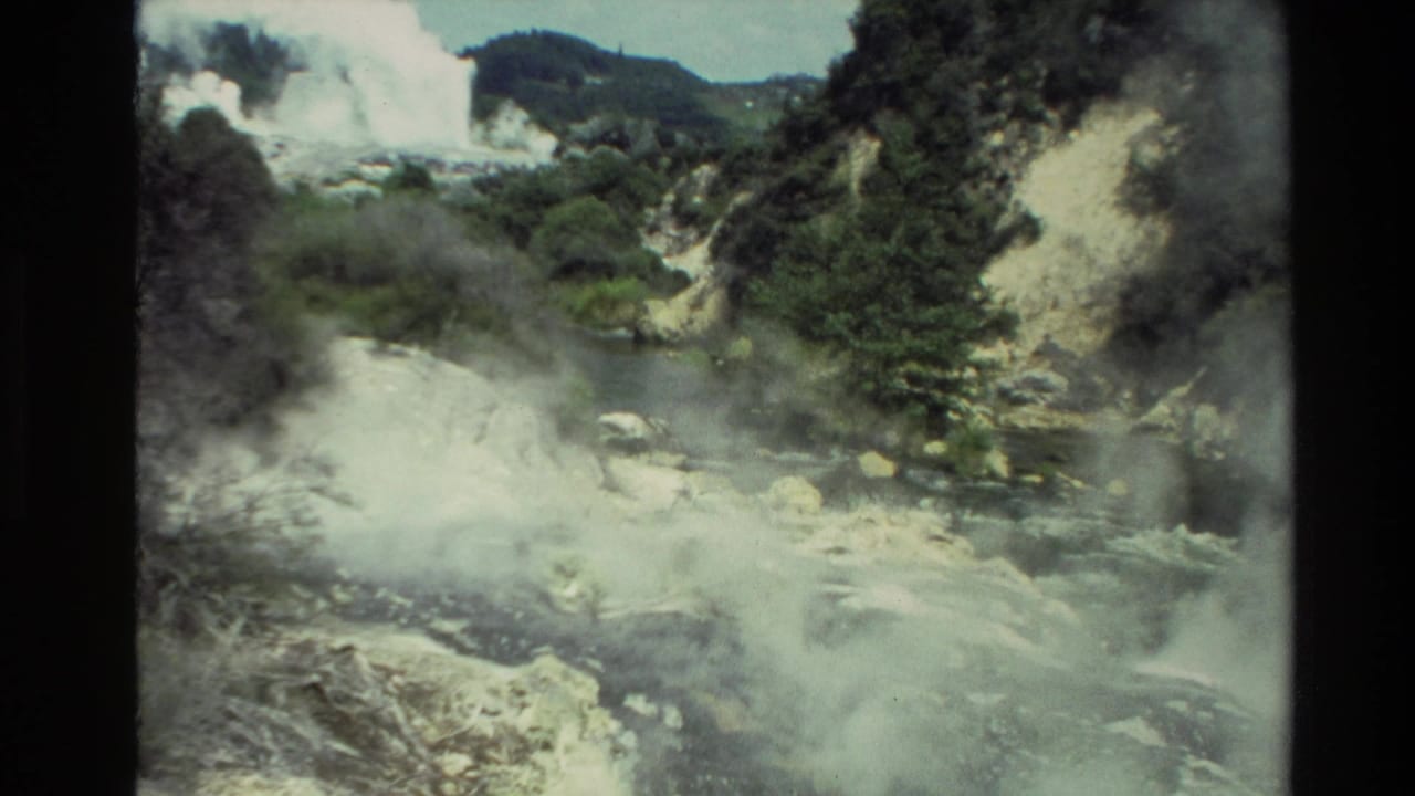 Steam rises from rocky geothermal ground near a stream and wooded hillside.