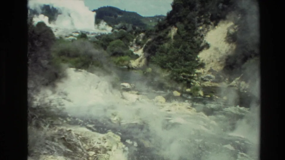 1981 New Zealand 8mm frame showing steam drifting over pale rocks, dark water, green vegetation, and a small distant cascade.