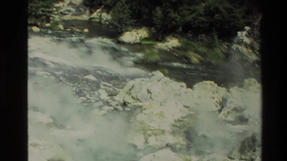 1981 New Zealand 8mm frame showing low steam over a rocky geothermal stream with bushes and shaded trees behind.