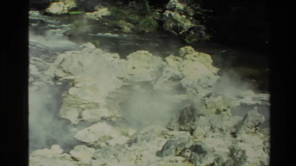 1981 New Zealand 8mm frame showing geothermal vapor around pale rocks, dark water, and vegetation at the edge of the stream.