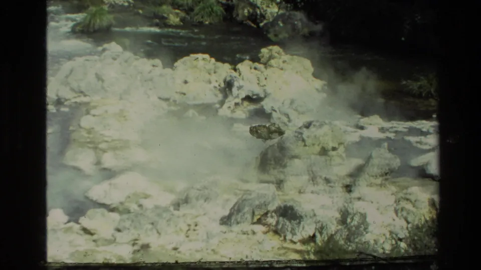 1981 New Zealand 8mm frame showing steam over rocky hot-spring water with trees and vegetation at the edge.