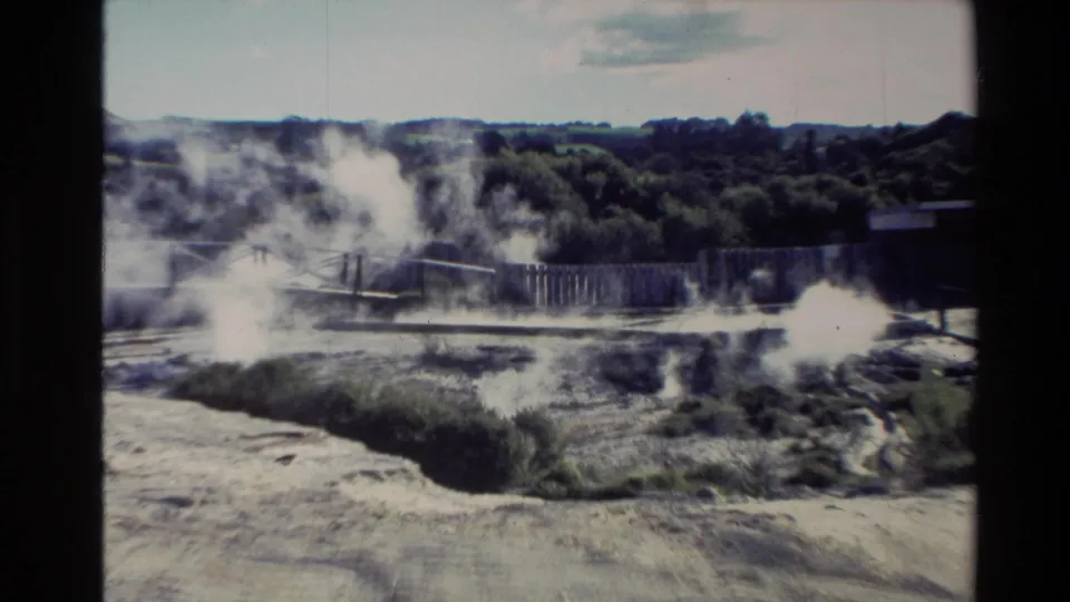 1981 New Zealand 8mm wide frame of a steaming geothermal area with fencing, pale earth, trees, and low hills.
