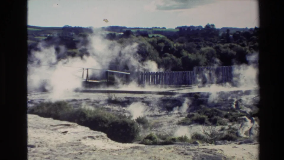 1981 New Zealand home-movie frame of steam vents, wooden fencing, rocky ground, and a treeline beyond.