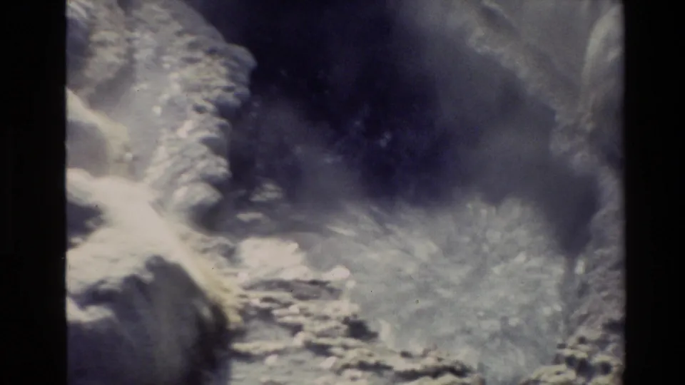 Close 8mm view of a New Zealand geothermal vent with steam, pale mineral deposits, and hot water among rocks.