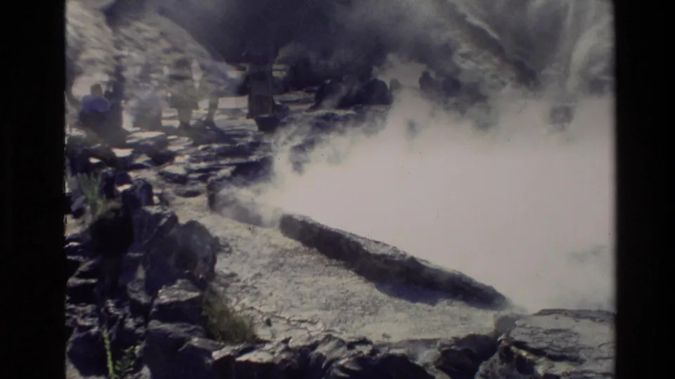 1981 New Zealand home-movie frame with tourists observing a steaming geothermal pool from a rocky viewing area.