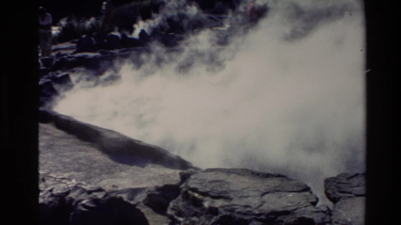 Steam rises over dark rocks and pale hot spring ground.