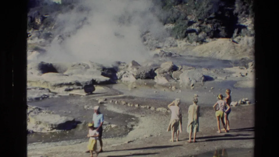 Visitors in casual 1981 clothing stand on pale ground near steaming geothermal pools and rocks in New Zealand.