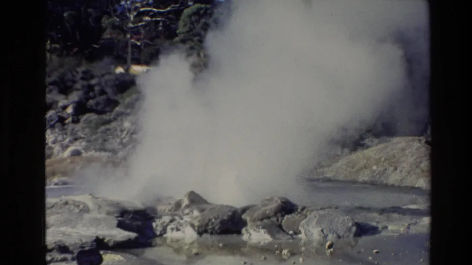 A large white steam cloud rises from rocky geothermal ground beside a pale pool in the New Zealand landscape.