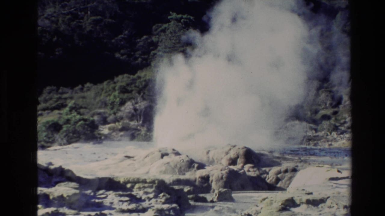 1981 New Zealand 8mm frame showing a white geyser plume rising from mineral-stained rocks with dark green trees in the background.
