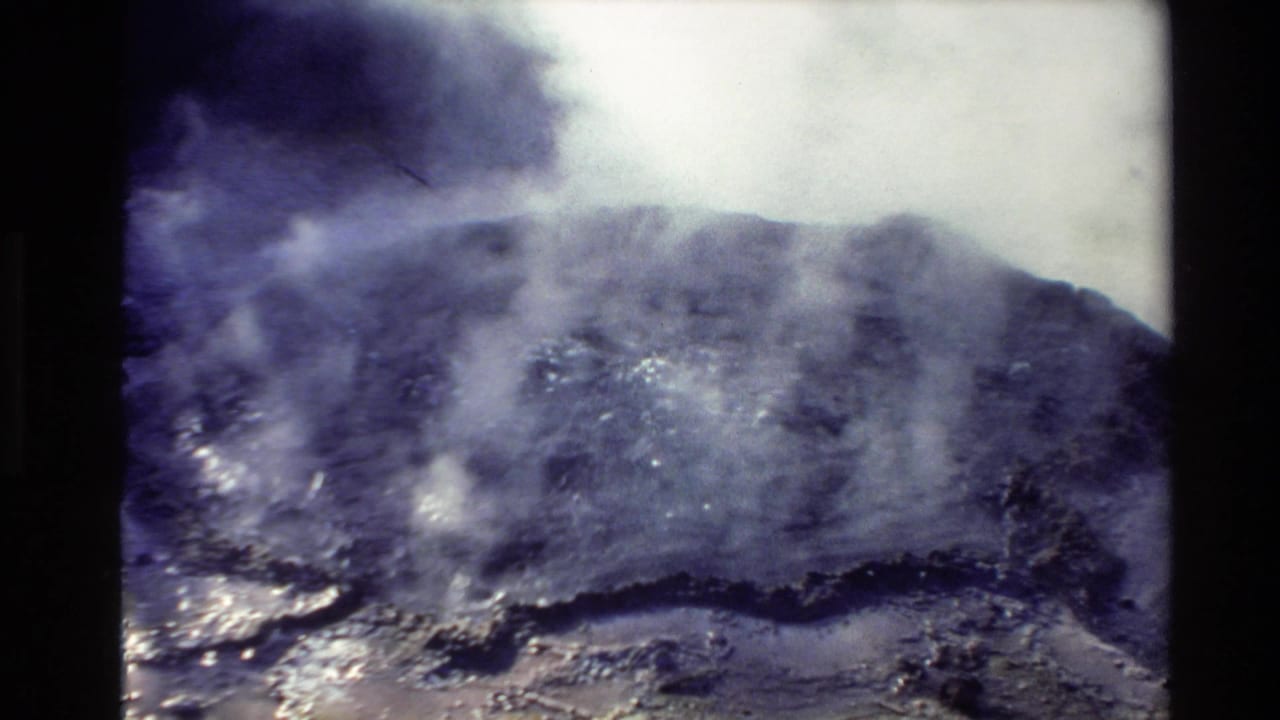 A grainy 8mm frame shows a dark, misty landscape or geothermal-looking surface, likely from the same New Zealand travel reel.