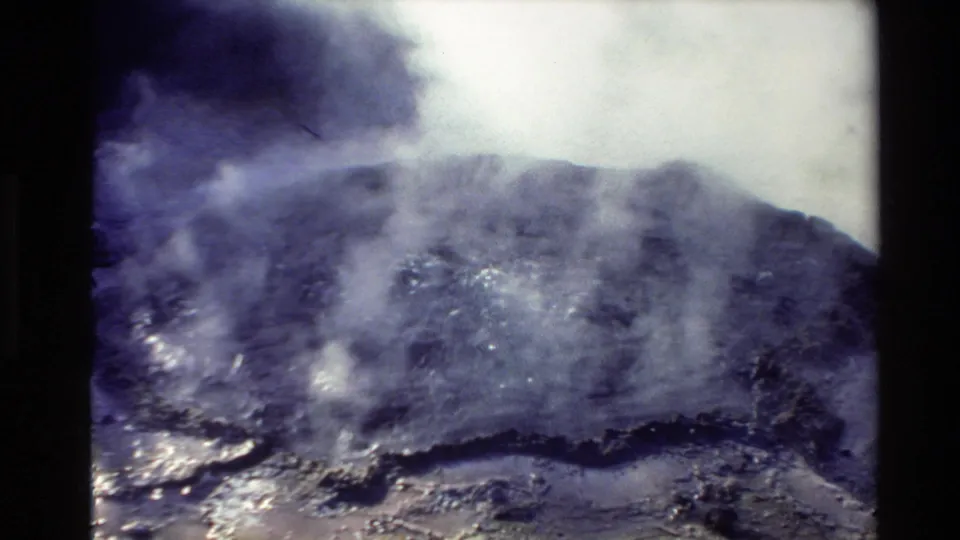 A grainy 8mm frame shows a dark, misty landscape or geothermal-looking surface, likely from the same New Zealand travel reel.