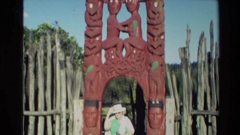 In 1981 New Zealand home-movie footage, a woman in a white hat appears beneath a red Maori carved wooden archway with fencing and trees behind.