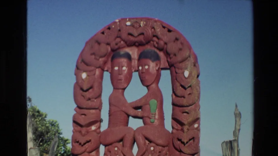 A red Maori carved archway fills the frame, showing two seated human figures, painted green accents, blue sky, and fence posts nearby.