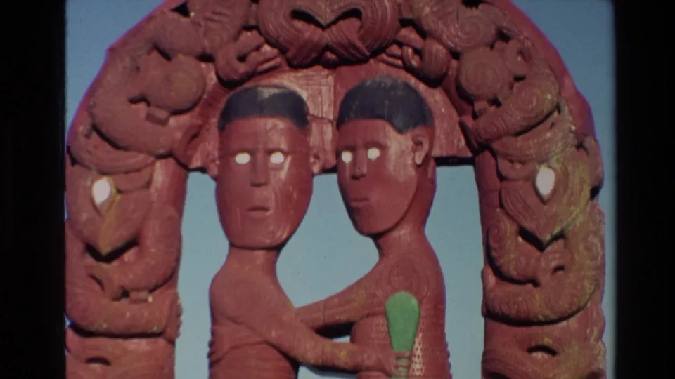 Close 8mm view of the red Maori carved archway, with two human figures, white eye details, green accents, and blue sky behind.