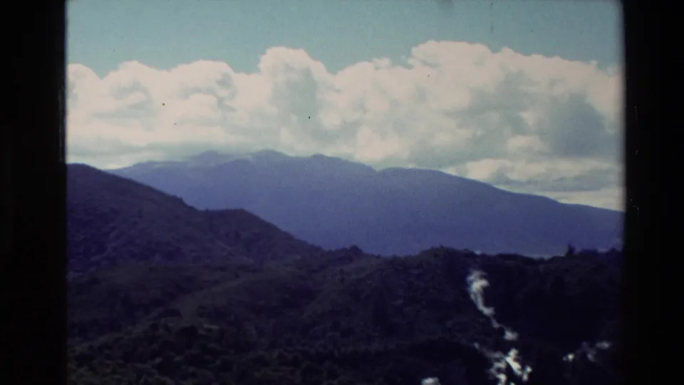 1981 New Zealand 8mm view across dark green hills toward distant mountains, with a bright river or waterfall line at right.