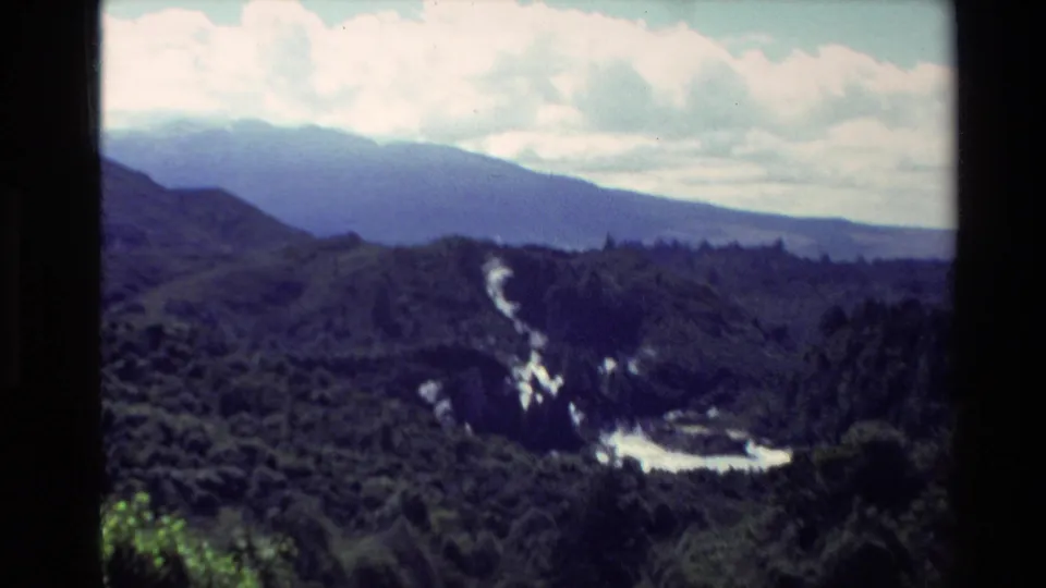 1981 New Zealand 8mm frame looking over a green valley toward hazy mountain slopes and a reflective river or waterfall area.