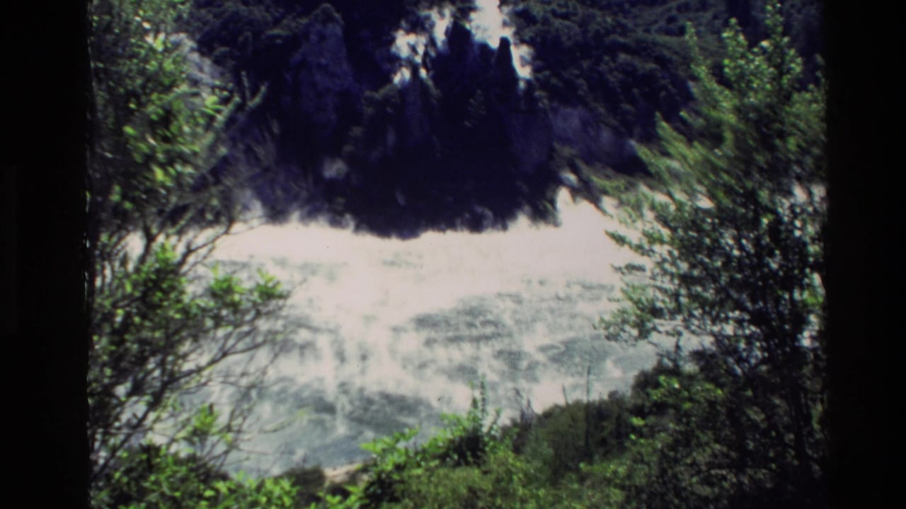 1981 New Zealand 8mm frame showing pale volcanic steam against a dark rocky hillside with green foliage in the foreground.